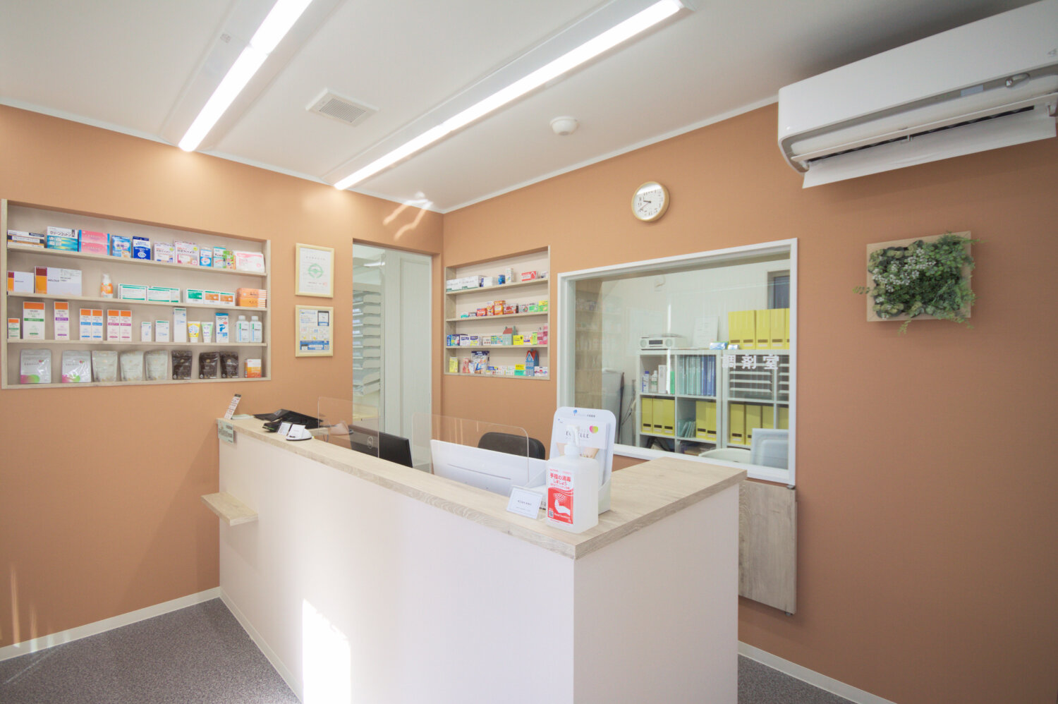 第２薬局 船橋店（千葉県船橋市）の受付カウンターとアクリルの仕切り、壁の棚、奥に調剤室のガラス窓（施工：有限会社Ai空間デザイン室）